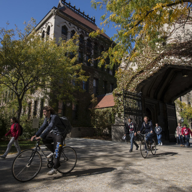 A student walks outside on a fall day while two cyclists ride through a leaf-covered archway on the UChicago campus.
