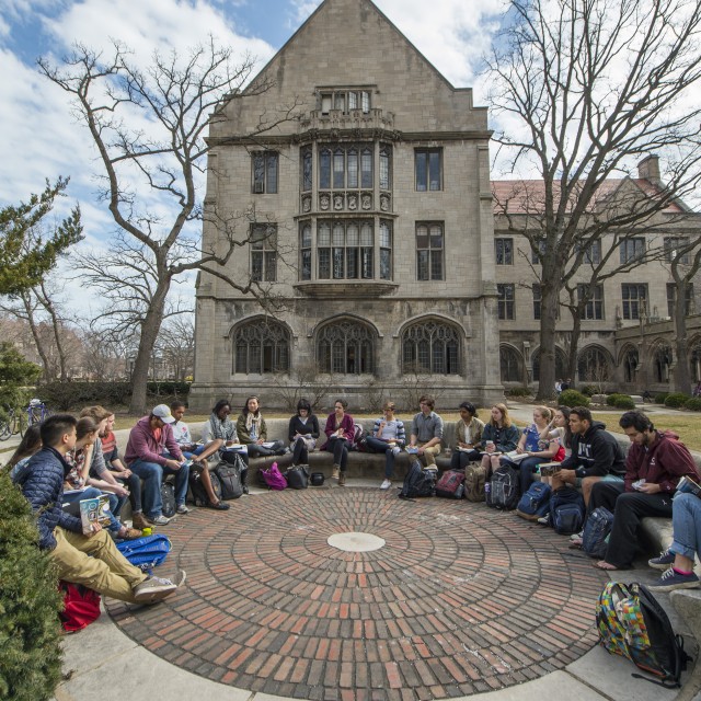 A class of students sits on a semi-circular stone bench on a cool day in fall, with leafless trees and a stone building framed behind the group.