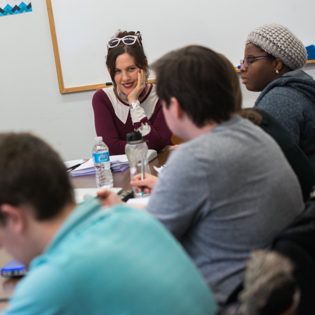 A smiling female-presenting professor sits at the head of a cozy table surrounded by students engaged in discussion.
