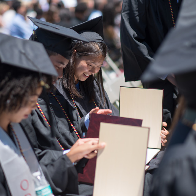 A row of seated students in cap and gowns look at their diplomas.