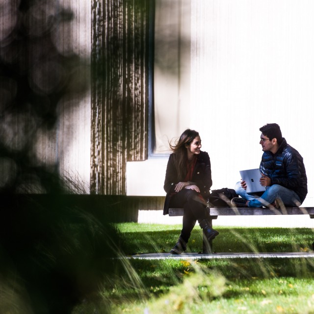 Two students wearing jackets sit on a bench outside talking on a sunny day.