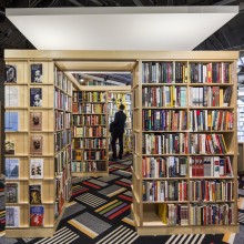 A male student stands amongst several shelves of books in a bookstore.