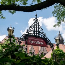 A banner that reads 'Congratulations from the College' is shown hanging from a large metal gate with bushes and trees in the foreground