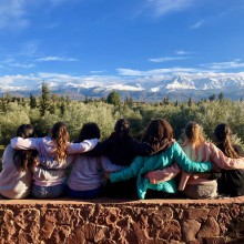 Viewed from behind. seven students sit on a stone wall, their arms slung around each other's shoulders, and look at the view of a forest with snow-capped mountains in the distance.