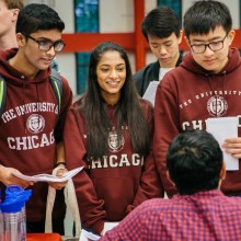 Two male students and a female student wearing UChicago maroon sweatshirts ask a male at a table a question.
