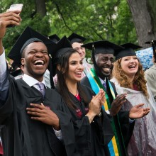 A group of students wearing caps and gowns clap and cheer, while one takes a selfie with a cell phone.