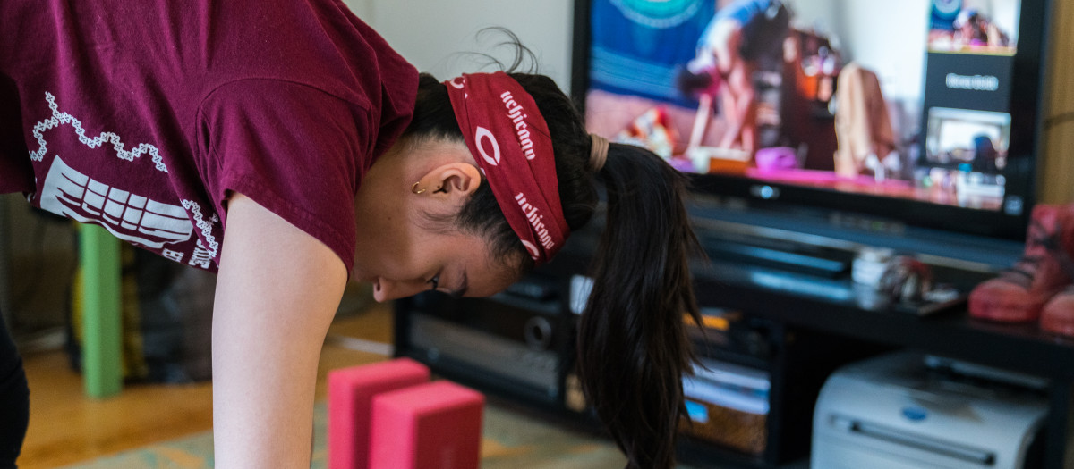 A student wearing a maroon tshirt and a University of Chicago bandana does a yoga pose while streaming a live yoga class to her TV.