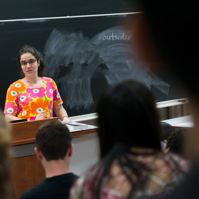 A female instructor stands in front of a blackboard while teaching a lecture class.
