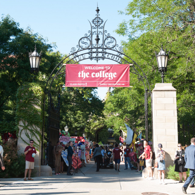 A crowd walks under an archway with a sign hanging that says "Welcome to the College"