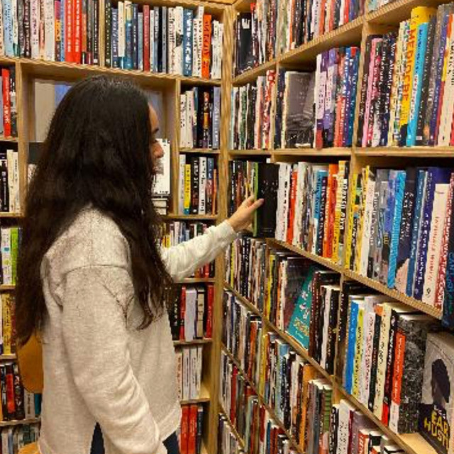 A woman stands in the corner of wooden library shelves filled with books touching a book.