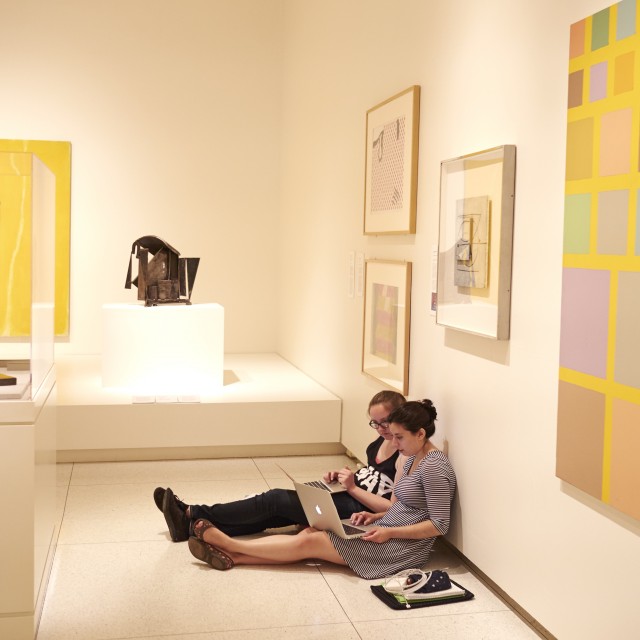 Two female-presenting students with laptops sit on the floor of an art gallery, their backs to the wall and legs outstretched.