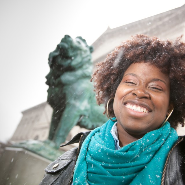 A close-up of a grinning female-appearing person in front of a bronze lion statue.