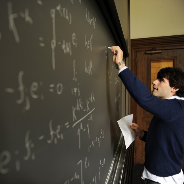 A male teacher writes equations on a blackboard in a classroom.