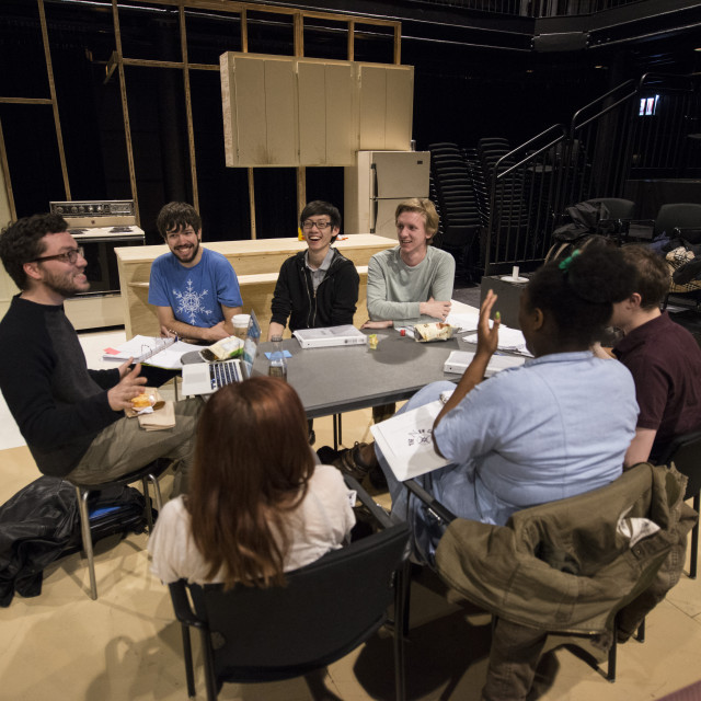 Theater students sit around a table in a sparse kitchen set, with an empty audience.