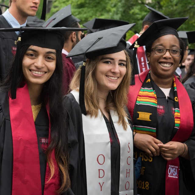 A group of students in graduation cap and gown on a sunny day. The focus is on three female-presenting people smiling at the camera.