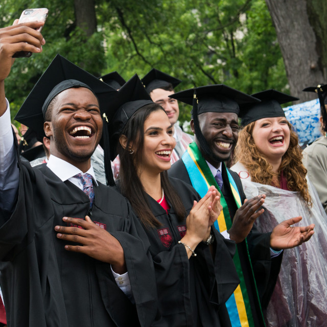 A group of students wearing caps and gowns clap and cheer, while one takes a selfie with a cell phone.