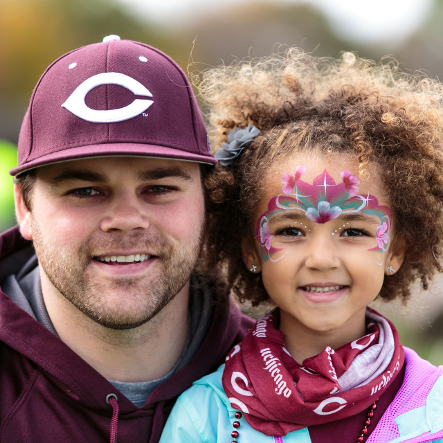 A young man in a maroon hat with the UChicago "C" logo on it posts with a little girl with floral facepaint on her face and a UChicago bandana around her neck.