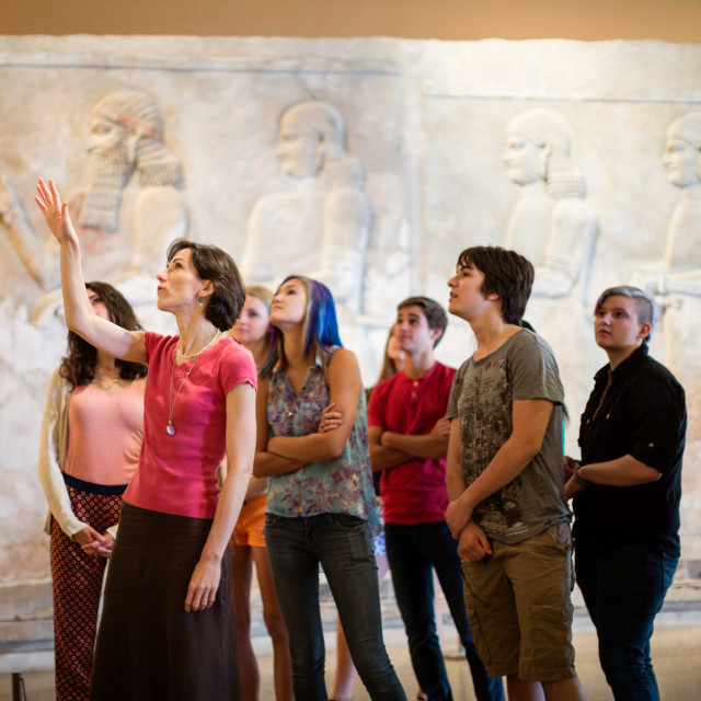 An instructor in a museum speaks to a group of students, pointing upward to something out of frame.
