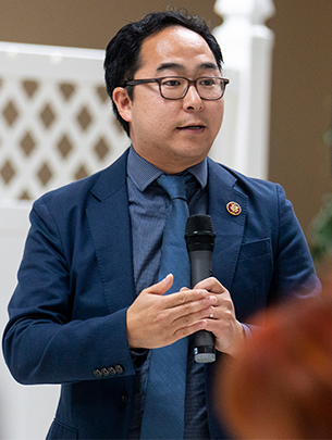 Rep. Andy Kim is shown speaking during a town hall meeting. A white picket fence is shown while he talks holding a microphone.