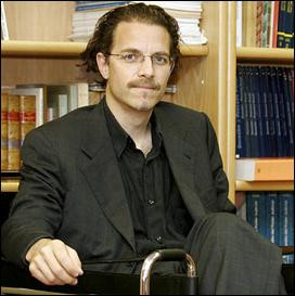 A male-presenting person sits in front of a bookshelf.