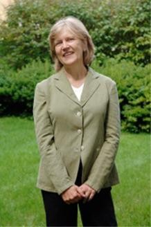 A female-presenting person stands on a grassy lawn in front of bushes with her hands clasped in front of her.