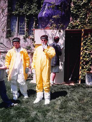 Two men stand wearing yellow suits stand in front of a breeder reactor. One smokes a cigarette.