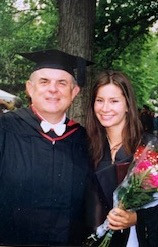 A man and woman pose wearing black graduation robes. The woman holds flowers.