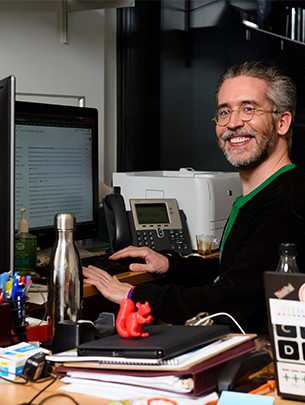 A profesor sits at a desk in front of two computer screens and a phone.