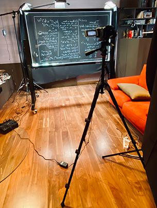 A camera is shown in a makeshift studio pointing at a clear marker board used to film lectures for a class.