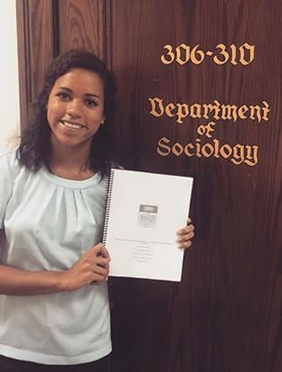McCammond holds a stack of papers aloft in front of a door that reads "Department of Sociology" in a gothic script.