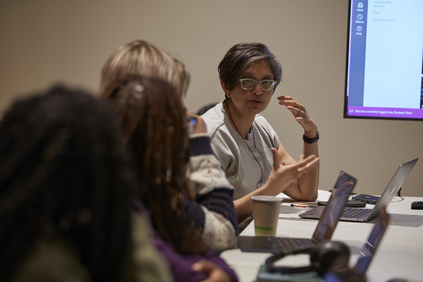 People in discussion around a conference table with laptops, coffee cups, and a screen displaying digital information.