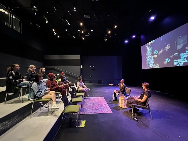 A group of people in a theater listens to two speakers on stage; a film is projected on a large screen.