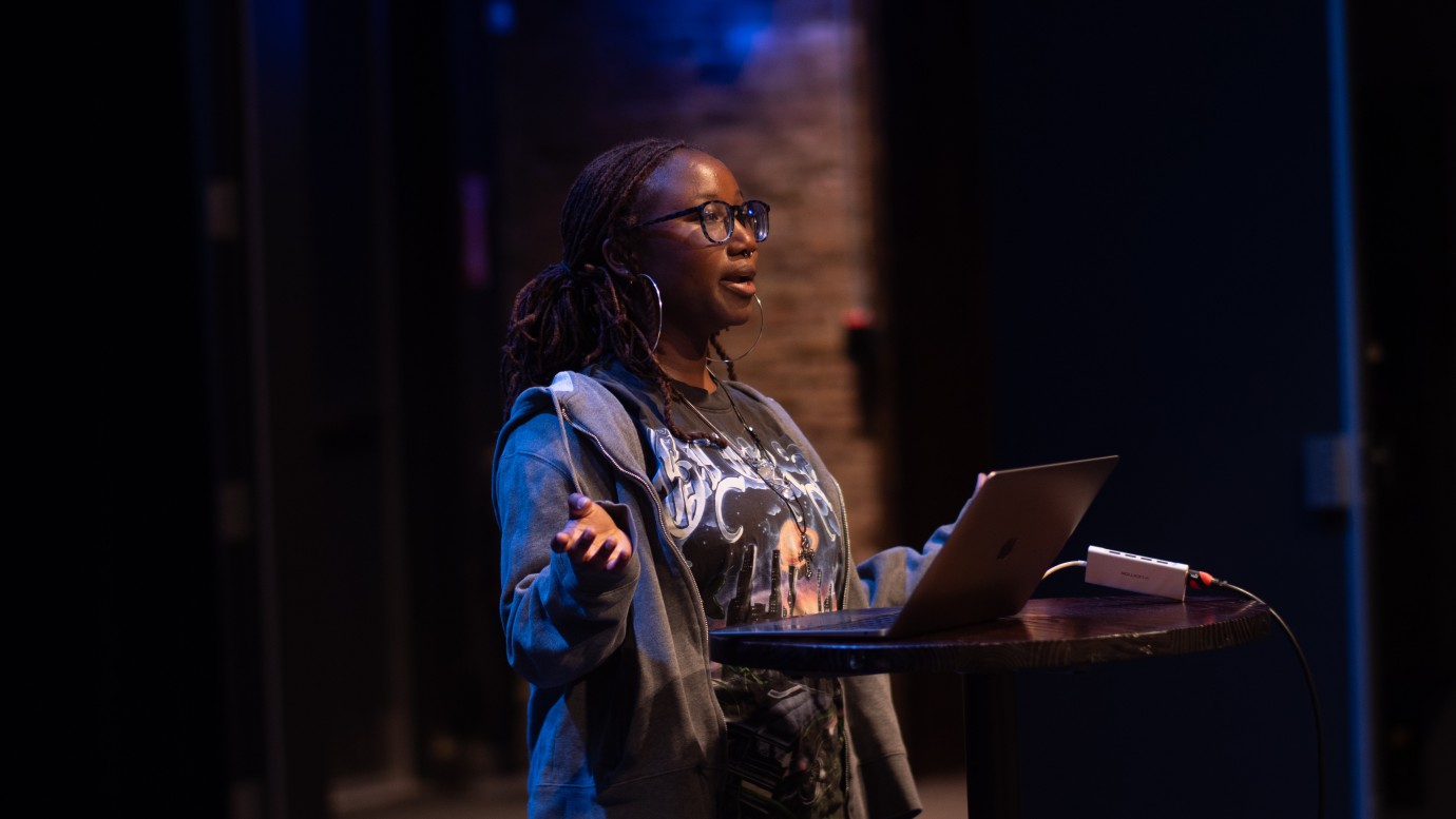 Person stands at a round table with a laptop in a dimly lit room with blue lighting and exposed brick wall.