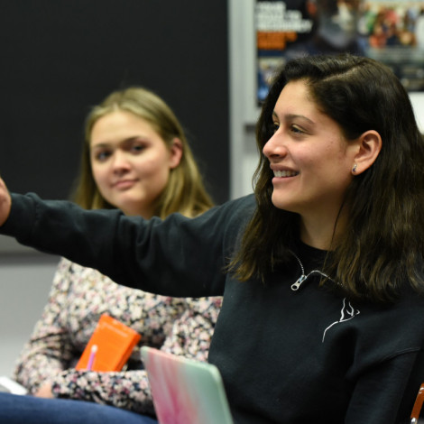 A student in a classroom raises her hand while another student in the background looks at her.
