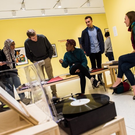 A group of four students sit listening to a record player with a professor in a yellow room in a museum.
